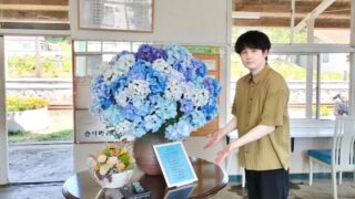 合川駅のアジサイと楽曲『アジサイ』の歌詞カードの前に立つ、音楽クリエイター日景健貴の写真