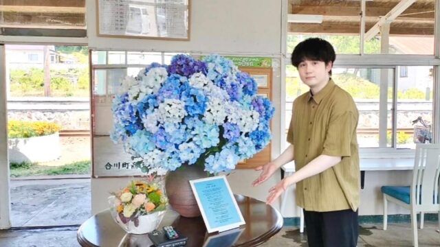 合川駅のアジサイと楽曲『アジサイ』の歌詞カードの前に立つ、音楽クリエイター日景健貴の写真
